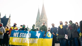 Zahlreiche Menschen haben sich während der Kundgebung gegen den Krieg in der Ukraine hinter dem Transparent "We stand with Ukraine" auf dem Domshof versammelt.