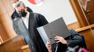 Der Angeklagte sitzt vor Prozessbeginn in einem Saal im Landgericht und bedeckt sein Gesicht mit einem Aktenordner, während sein Anwalt Detlev Otto Binder neben ihm steht. Der Mann soll im Oktober 2021 in Delmenhorst zunächst in einer Bar einen vermeintlichen Nebenbuhler erstochen haben.