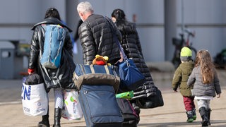 Drei Erwachsene und zwei Kinder sind von hinten zu sehen. Die Erwachsenen ziehen Koffer und tragen Taschen.