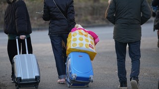 Drei Erwachsene und zwei Kinder sind von hinten zu sehen. Die Erwachsenen ziehen Koffer und tragen Taschen.