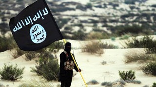 Ein maskierter Mann mit einer Fahne des sogenannten Islamischen Staates steht vor einer Wüstenkulisse.