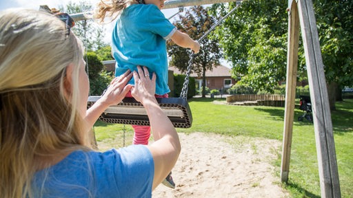 Eine Frau gibt ihrer Tochter auf einem Spielplatz Anschwung beim Schaukeln