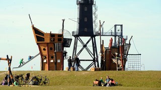 Menschen sitzen vor einem Klettergerüst und einem Leuchtturm auf dem Deich.