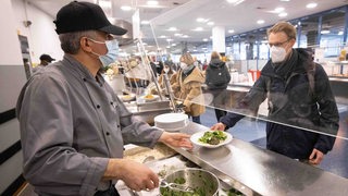 Ein Mitarbeiter einer Uni-Mensa gibt ein Essen an einen Studierenden aus. Beide tragen einen Mund-Nasen-Schutz