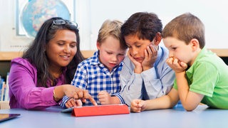 Eine Lehrerin mit Grundschülern an einem Tablet. Verschiedene Nationalitäten und Hautfarben