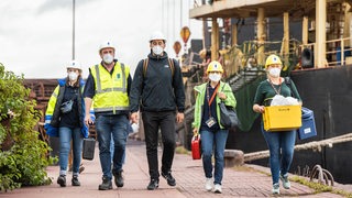 Mehrere Menschen mit Helmen, Warnwesten und Ausrüstung laufen neben einem Schiff.