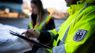 Beamtinnen und Beamte vom Zoll kontrollieren auf dem Gelände eines Logistikunternehmens den Inhalt eines importierten Containers.