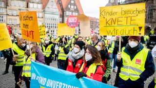 Mitarbeiter kommunaler Kitas und sozialer Dienste stehen während einer Kundgebung mit Plakaten auf dem Marktplatz, u.a. mit einem Plakat "Kinder brauchen Knete, wir auch!". 