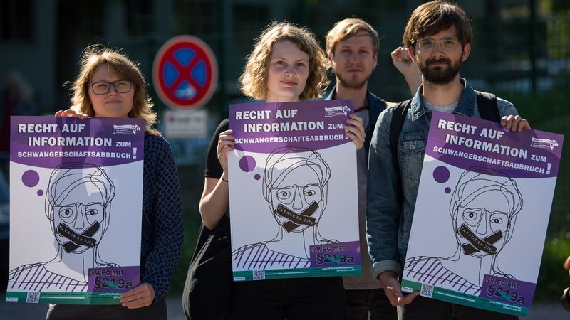 Frauen und Männer mit Plakaten in den Händen. Auf den Plakaten ein gezeichnetes Gesicht mit zugeklebten Mund und der Aufschrift "Recht auf Information zum Schwangerschaftsabbruch"