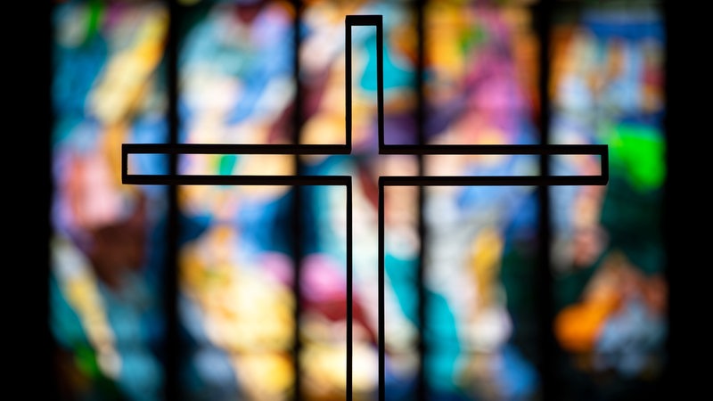 Ein Kreuz zeichnet sich vor einem bunten Glasfenster ab 