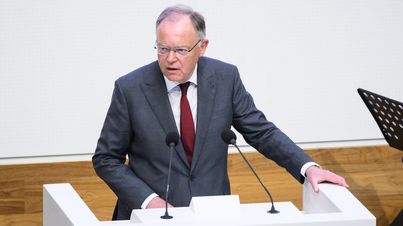 Ein Mann mit grauen Haaren, Brille und grauem Anzug sowie roter Krawatte steht an einem Rednerpult mit der Aufschrift "Niedersächsischer Landtag" und spricht. 