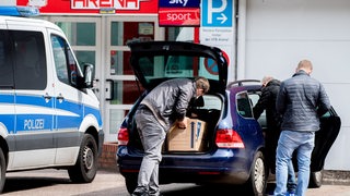 Ermittlungsbeamte der Polizei verstauen vor einer Sporthalle mehrere Umzugskartons in Einsatzfahrzeugen. 