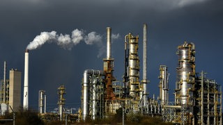 Bild einer Ölraffinerie. Über der Raffinerie sind dunkle Wolken zu sehen. 