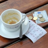 Münzen, ein Geldschein und eine Rechnung liegen neben einer leeren Kaffeetasse auf einem Tisch.