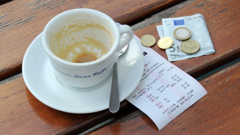 Münzen, ein Geldschein und eine Rechnung liegen neben einer leeren Kaffeetasse auf einem Tisch.