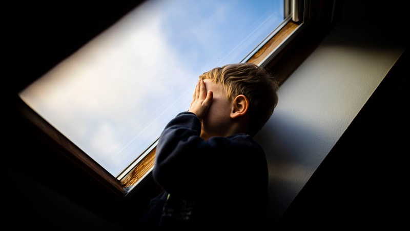 Ein kleiner Junge steht am Fenster und hält sich die Hände vor sein Gesicht.
