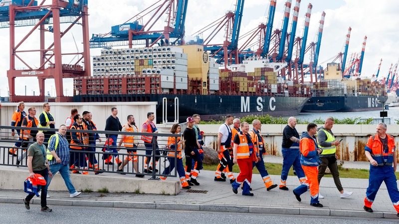 Hafenarbeiter gehen am Hamburger Hafen zu einer Streikversammlung, im Hintergrund wird ein Containerschiff beladen.