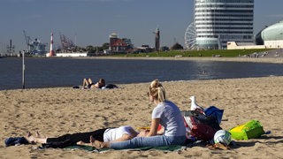 Menschen sitzen an einem Sandstrand vor einer Kulisse aus Wasser und Gebäuden.