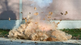 Ein Blitzknallsatz explodiert. Eine Rauchwolke steigt auf, eine rote Stichflamme ist zu sehen.