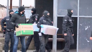 Polizisten tragen Kartons aus einem Gebäude.
