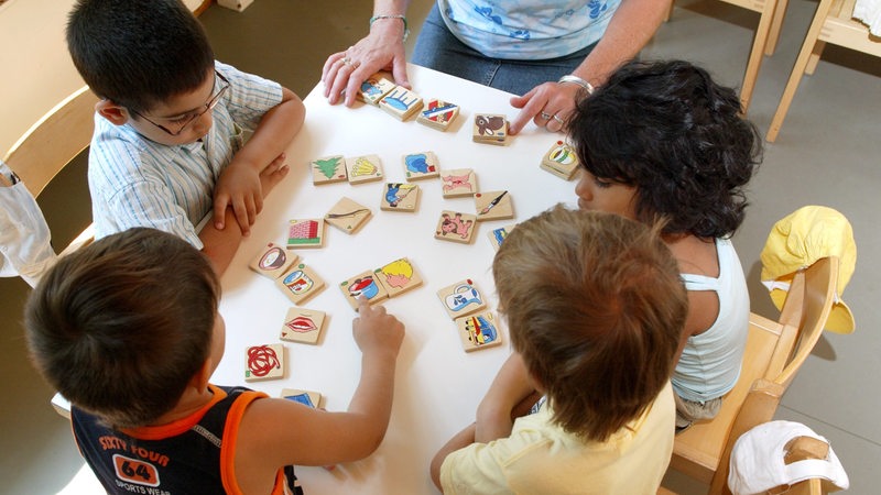 Mit Hilfe eines Spiels vermittelt eine Erzieherin vier Kindern an einem Tisch Wörter und Sprache.