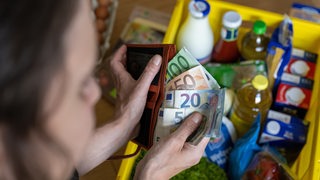 Eine Frau hält ein Portemaine und Geldscheine in den Händen. Darunter steht eine Kiste mit Lebensmitteln von einem Einkaur.