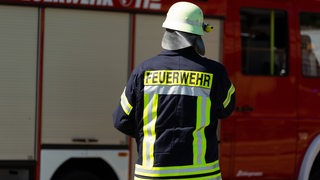 Ein Feuerwehrmann mit Sicherheitshelm und dem Aufdruck Feuerwehr auf seiner Einsatzkleidung. Im Hintergrund ist ein Einsatzfahrzeug, Feuerwehrwagen mit Schriftzug und Blaulicht zu sehen zu sehen. 