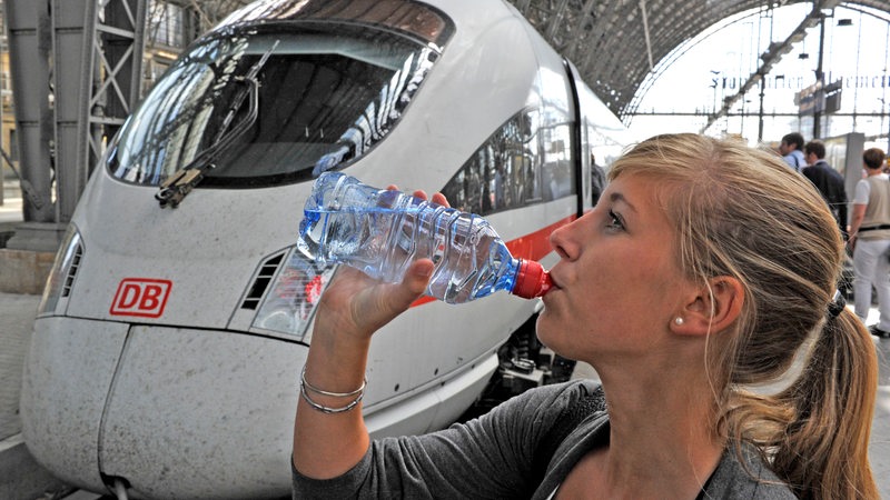 Eine Frau steht auf einem Bahnsteig und trinkt Wasser. Im Hintergrund steht ein ICE.