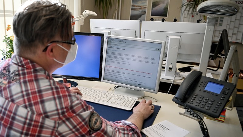 Ein Mann sitzt mit Maske an seinem Schreibtisch.