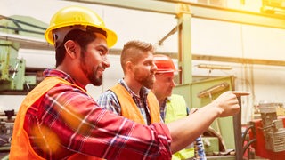 Drei Männer mit Warnweste und Schutzhelm stehen in einer Fabrik