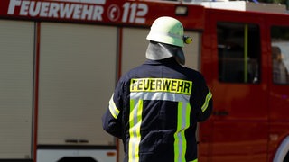 Ein Feuerwehrmann steht vor einem Feuerwehrwagen. Er dreht den Rücken zur Kamera.
