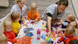 Einen Kindergartengruppe sitzt zusammen mit zwei jungen Freiwilligen im Sozialen Jahr an einem Maltisch