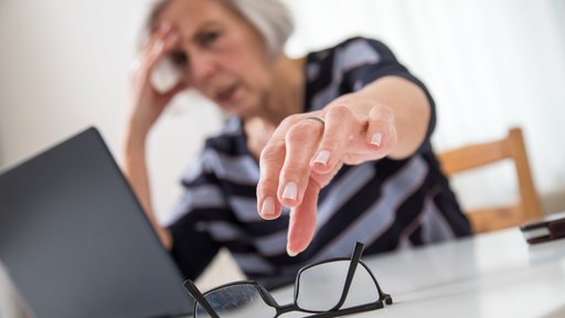 Eine ältere Frau sitzt am Laptop und greift nach ihrer Brille.