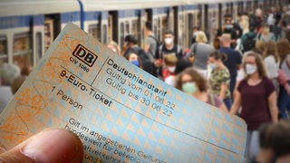Eine Hand hält ein 9-Euro-Ticket. Dahinter ist ein Bahnsteig zu sehen, auf dem viele Menschen sind und in einen Zug einsteigen. 