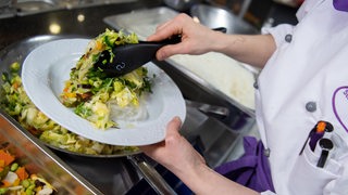 Eine Frau serviert in einer Mensa ein veganes Gericht mit Glasnudeln und Chinakohl. 