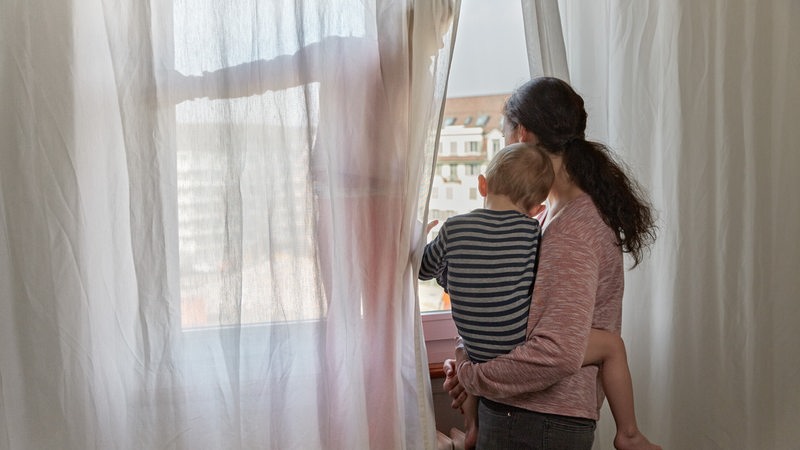Eine Mutter schaut mit Kind auf dem Arm aus dem Fenster.