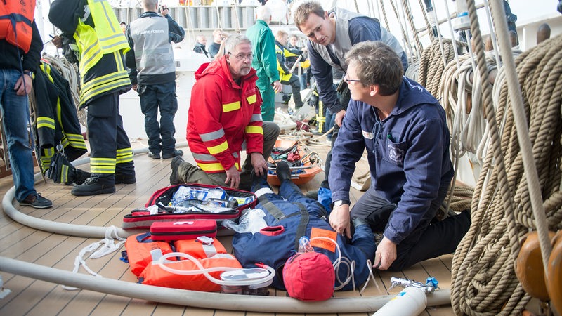 Mehrere Rettungskräfte knien neben einer Puppe auf einem Schiff.
