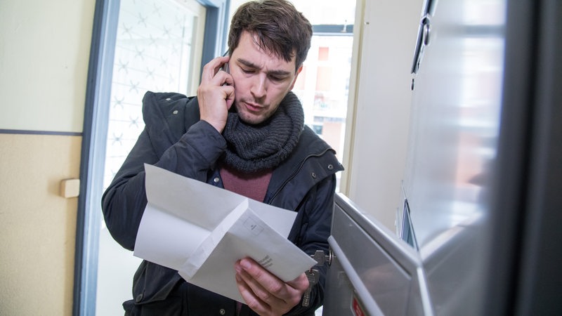 Ein Mann liest in einem Wohnhaus in Hamburg vor seinem Briefkasten einen Brief und telefoniert währenddessen mit seinem Smartphone.