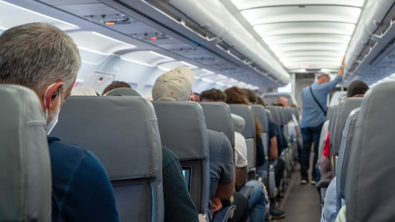 Passagiere sitzen in Flugzeug auf ihren Sitzen. Ein Mann steht im Gang.