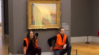 Eine Frau und ein Mann in orangener Weste kleben mit einer Hand an der Wand in einem Museum. An der Wand hängt ein Gemälde, das mit Farbe beschmiert wurde. 