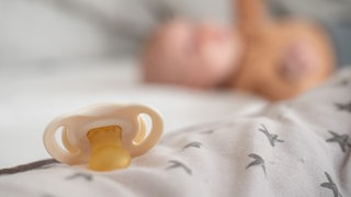 Ein Baby-Schnuller auf einem Tuch, im Hintergrund ein Baby.