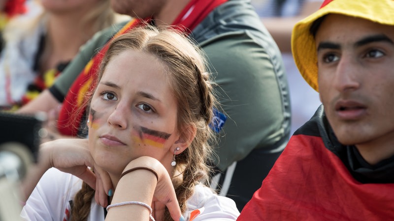 Junge Frau mit Deutschland-Flagge aufgemalt auf dem Gesicht guckt enttäuscht. Daneben andere Fußballfans.