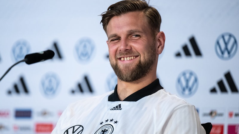 Werder-Stürmer Niclas Füllkrug grinst bei einer WM-Pressekonferenz so breit, dass seine markante Zahnlücke aufblitzt.