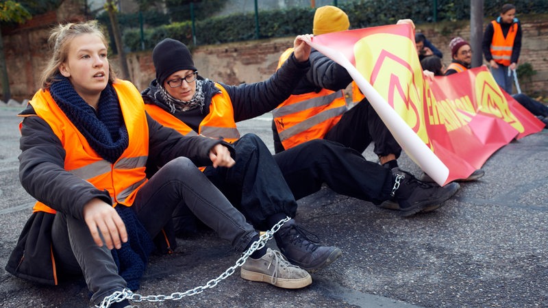 Klima-Aktivisten sitzen mit Ketten an den Füßen auf einer Straße.