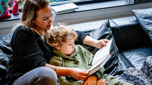 Eine Frau liest einem kleinen Jungen auf einem Sofa ein Buch vor.
