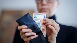 Eine Frau mit Brille guckt in eine Geldbörse und hält 10 Euro in der Hand.