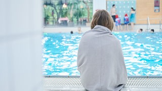 Ein Mädchen sitzt am Rand eines Schwimmbeckens und ist mit einem Handtuch bedeckt.