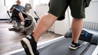 Ein Mann trainiert an einem Sportgerät, um seine Ausdauer nach einer Coronainfektion zu stärken, während ein anderer Patient auf einem Laufband trainiert.