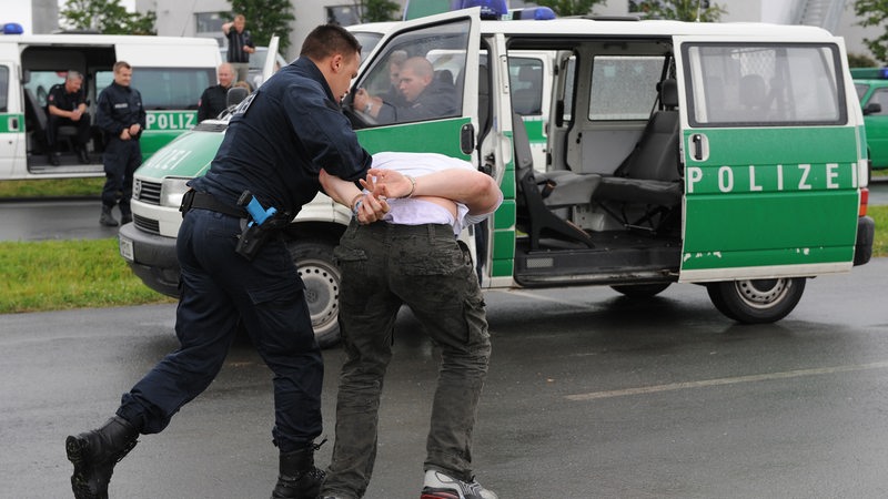 Ein Polizist führt einen Mann in Handschellen zu einem Polizei-Fahrzeug