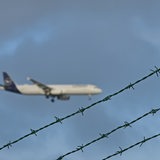 Ein Flugzeug ist am Himmel zu sehen. Im Vordergrund ist Stacheldraht zu sehen.
